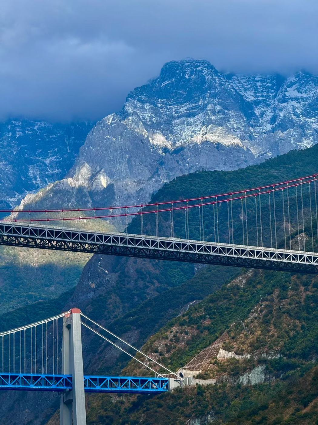 Two suspension bridges across the valley with the snow-streaked Jade Dragon mountain behind under heavy cloud