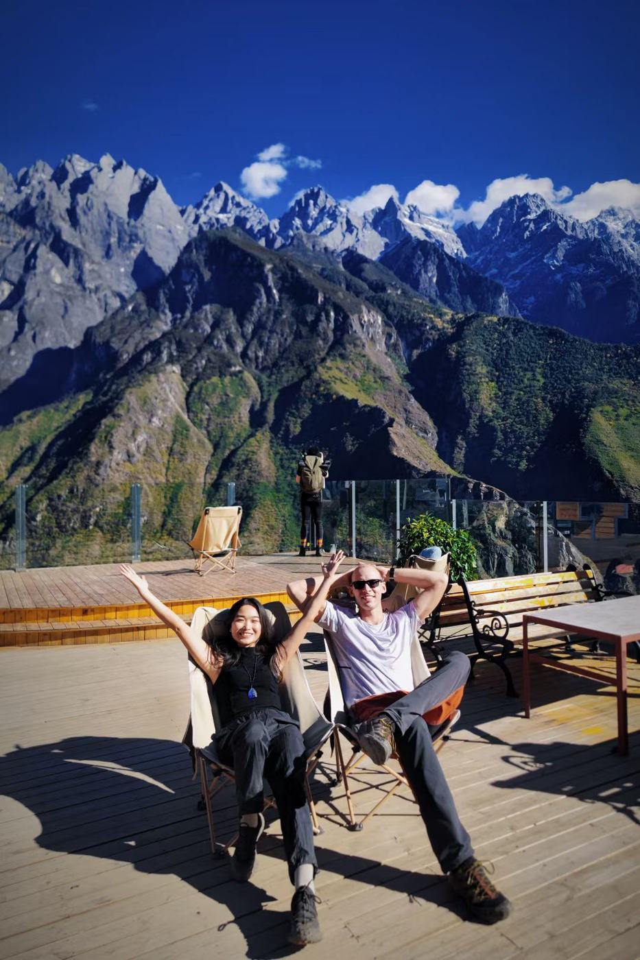 Two hikers relaxing in folding chairs on a wooden terrace, with the Jade Dragon range behind