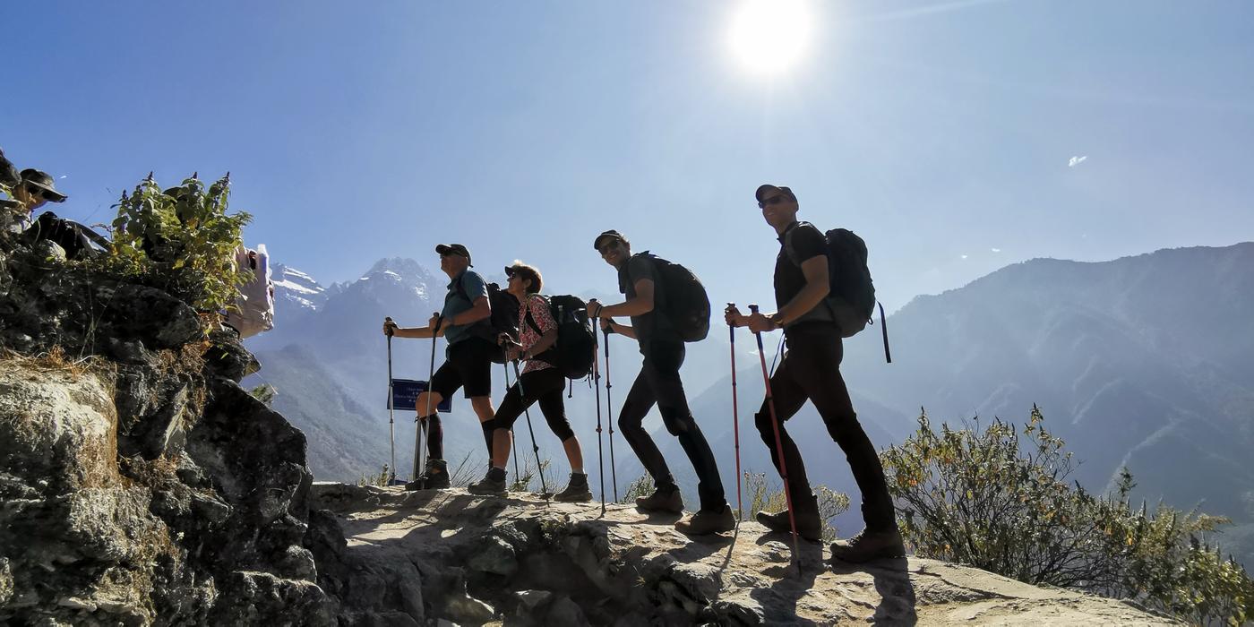 Four hikers with trekking poles on a rocky ridge against the sun, with snow peaks behind them