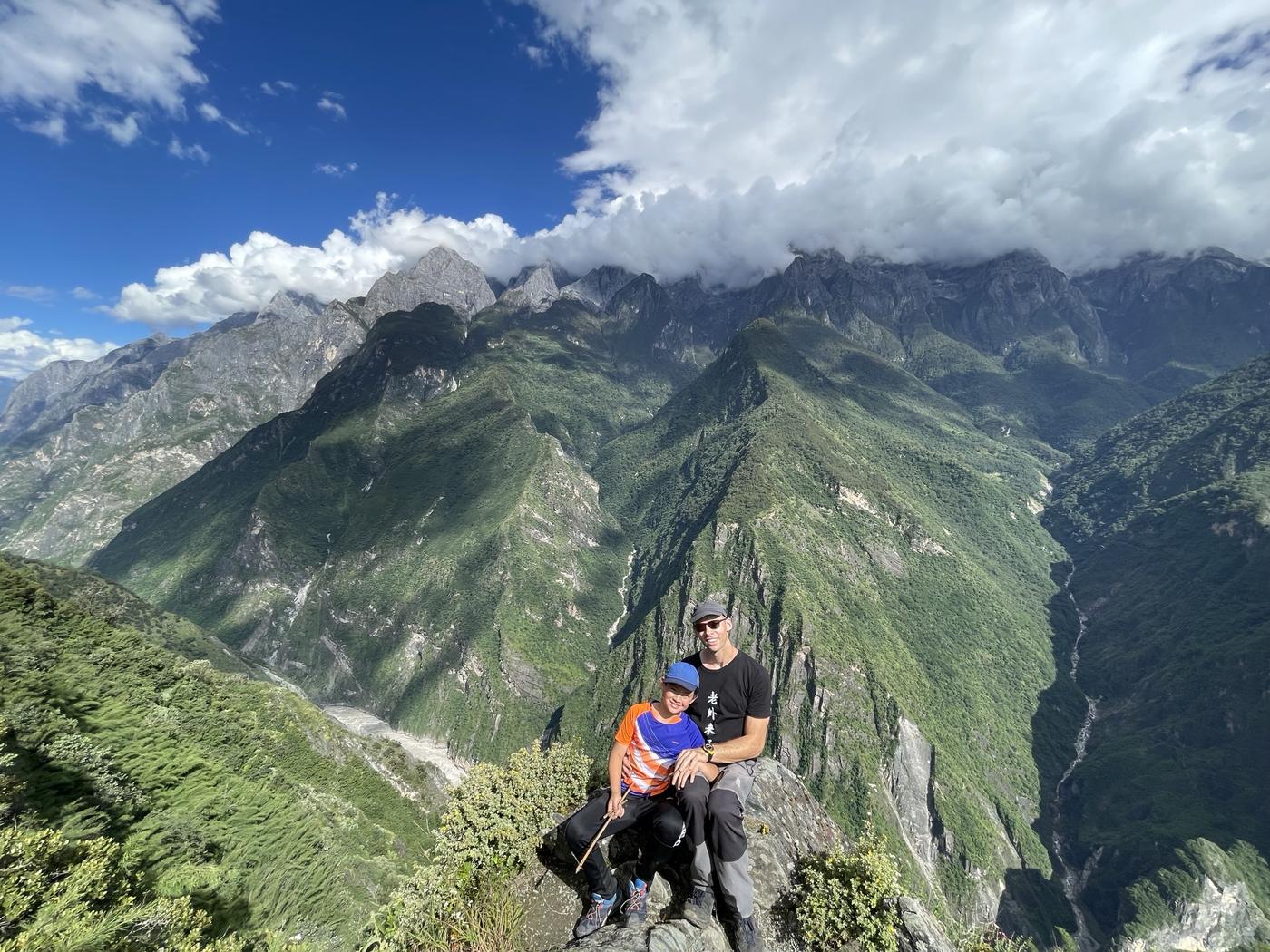 Two hikers sitting on a rock above the gorge, with the green slopes and the river far below and cloud over the peaks